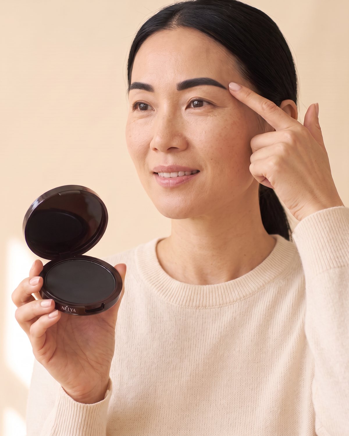 O femeie cu sprâncenele conturate își atinge sprânceana, cu cutiuța Nélya în mână